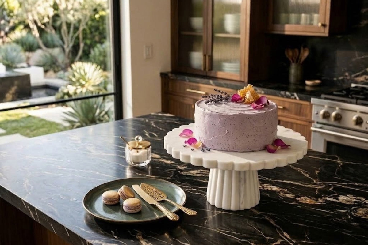 White marble cake stand with a scalloped rim carrying a lavender-frosted cake on a dark marble island