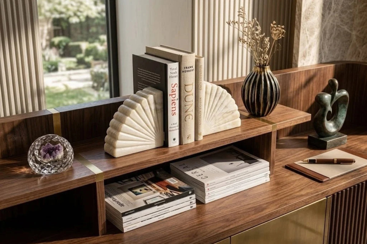 Ivory onyx bookends with fanned ridges holding hardbacks upright on a walnut desk shelf by a garden window