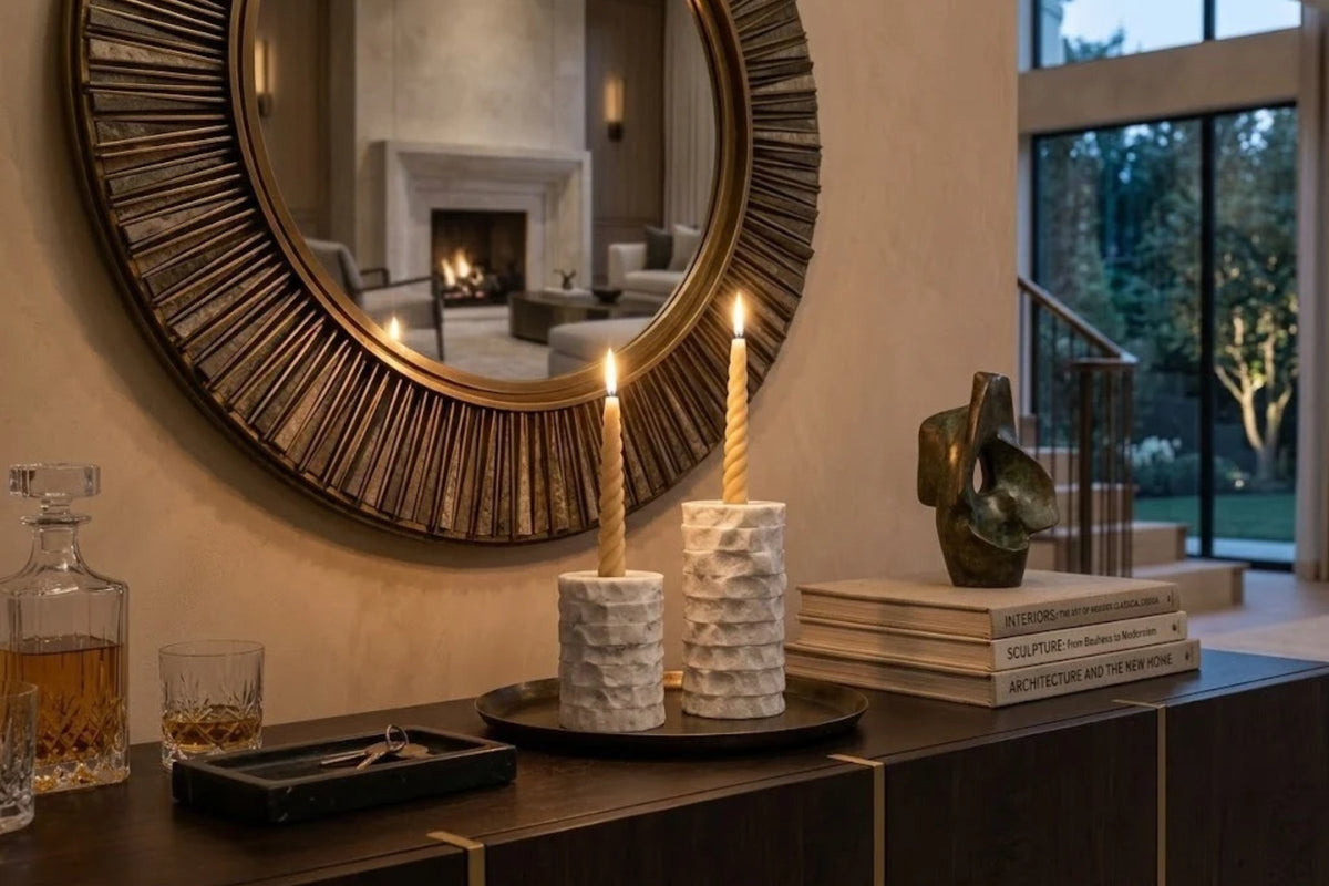 White marble candle holders chiseled in rough bands, two lit tapers on a dark tray on a console table