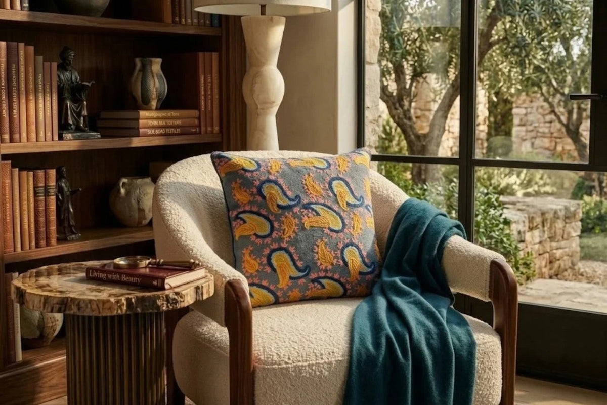 Slate blue paisley pillow in wool embroidery on a bouclé chair beside bookshelves and a stone side table