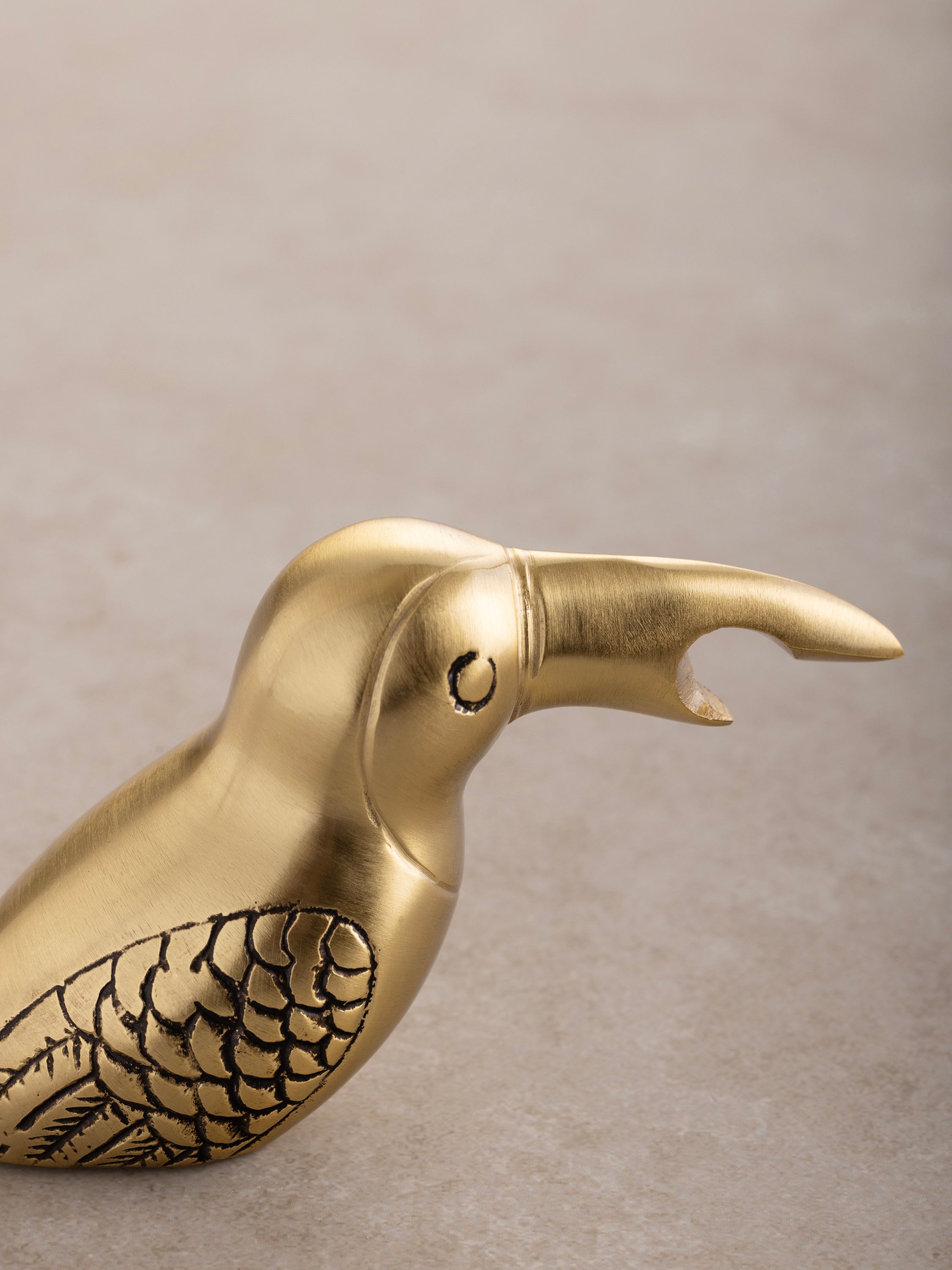 Close-up of the Goldie opener showing its carved feather detailing, elongated beak form, and polished brass surface.