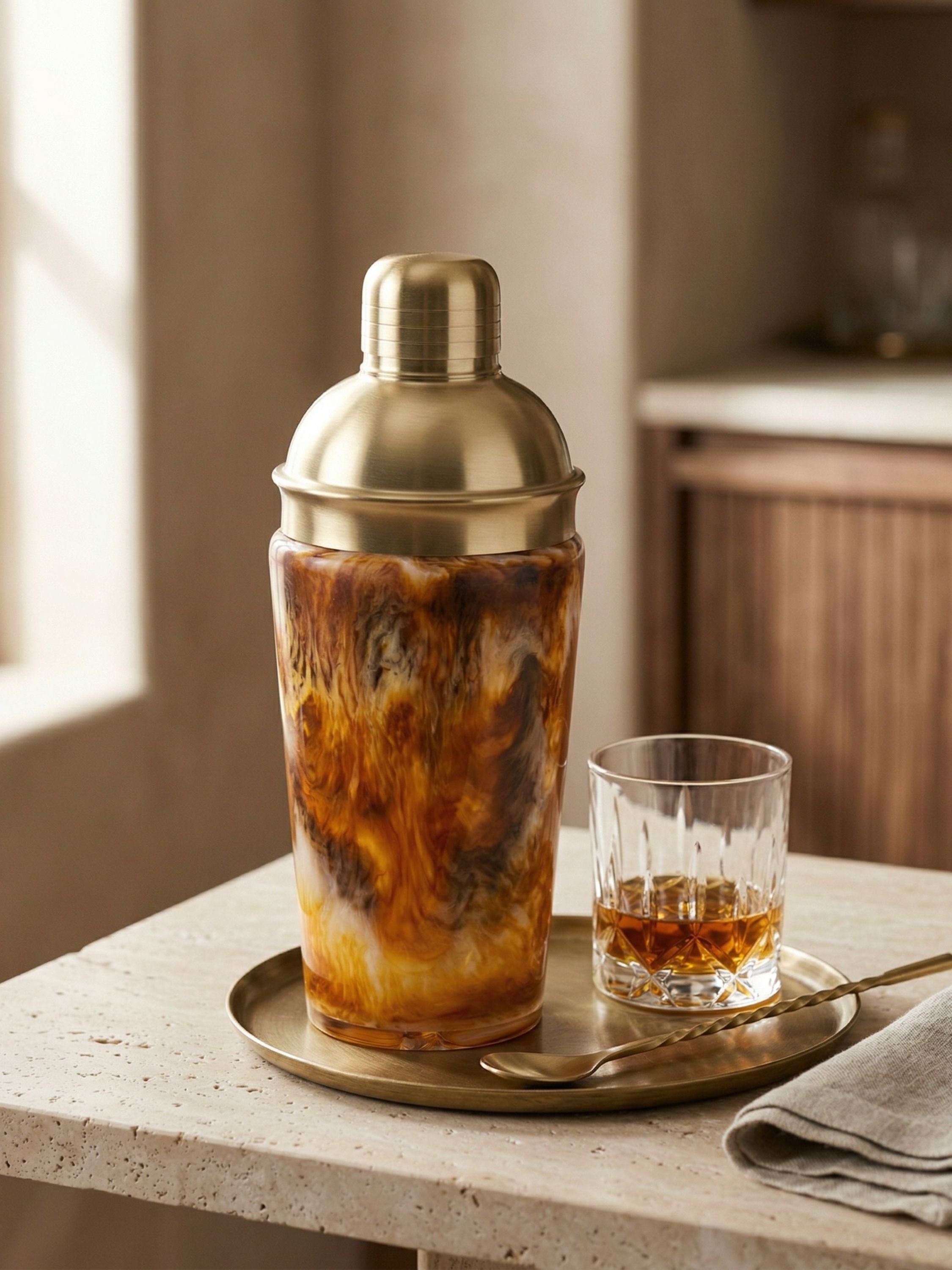 Cocktail shaker with a drink on a tray next to a glass on a countertop.