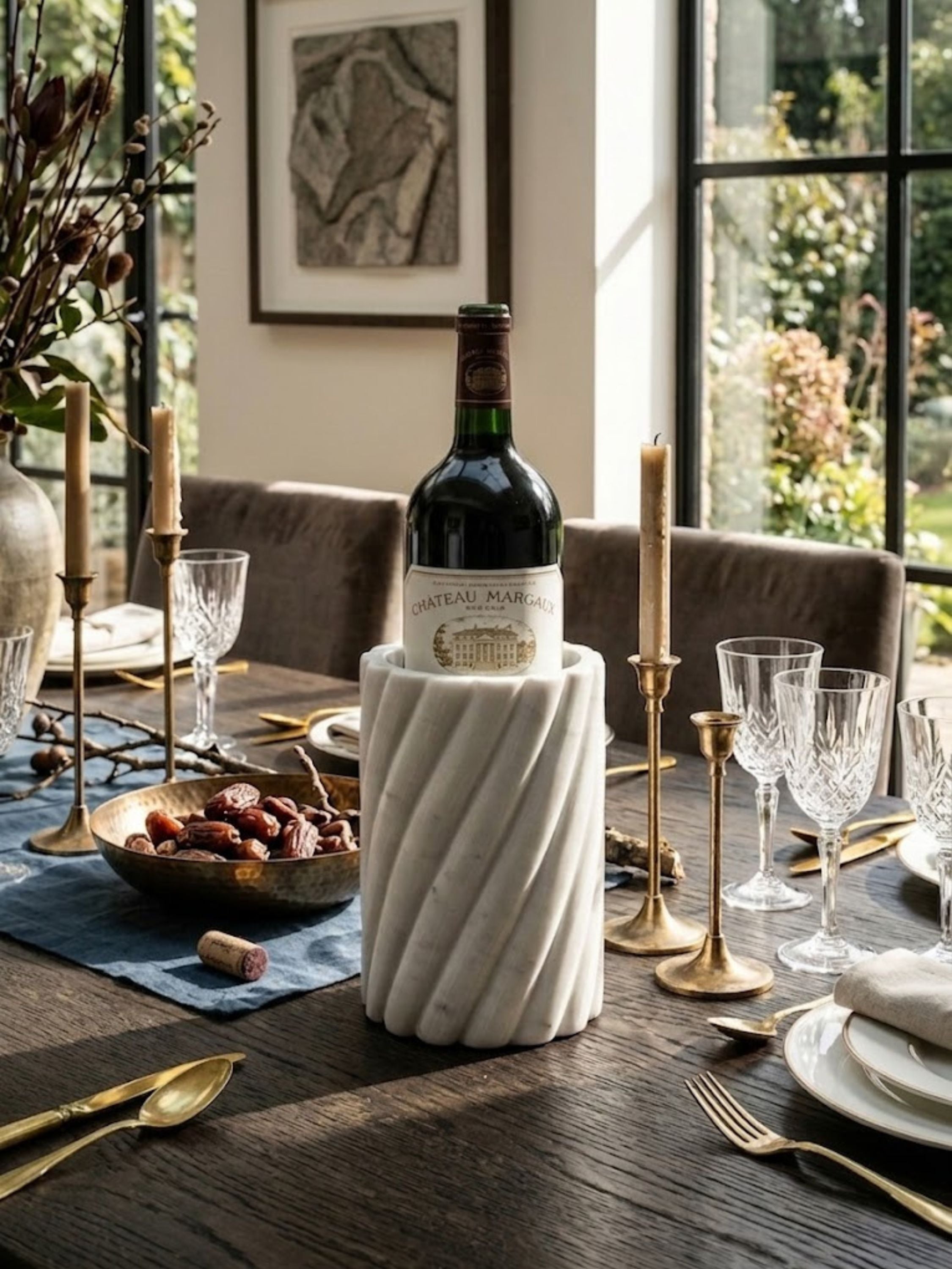 Sculptural white marble wine cooler with fluted form displayed on a dining table, showcasing a wine bottle among crystal stemware and brass candleholders.