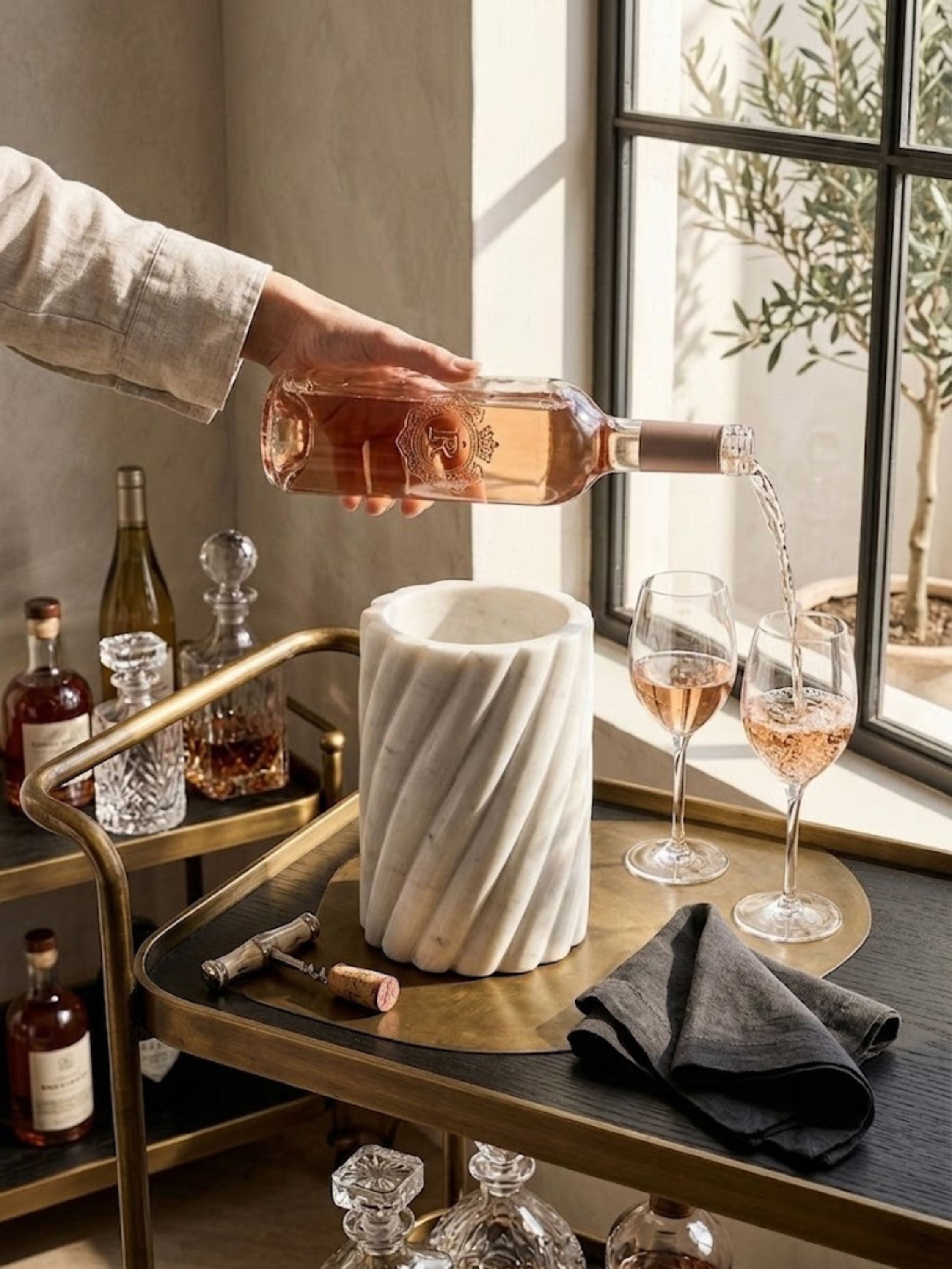 Taraz white onyx wine cooler with sculpted spiral fluting styled on a brass bar cart, paired with crystal glassware and rosé beside a sunlit window.