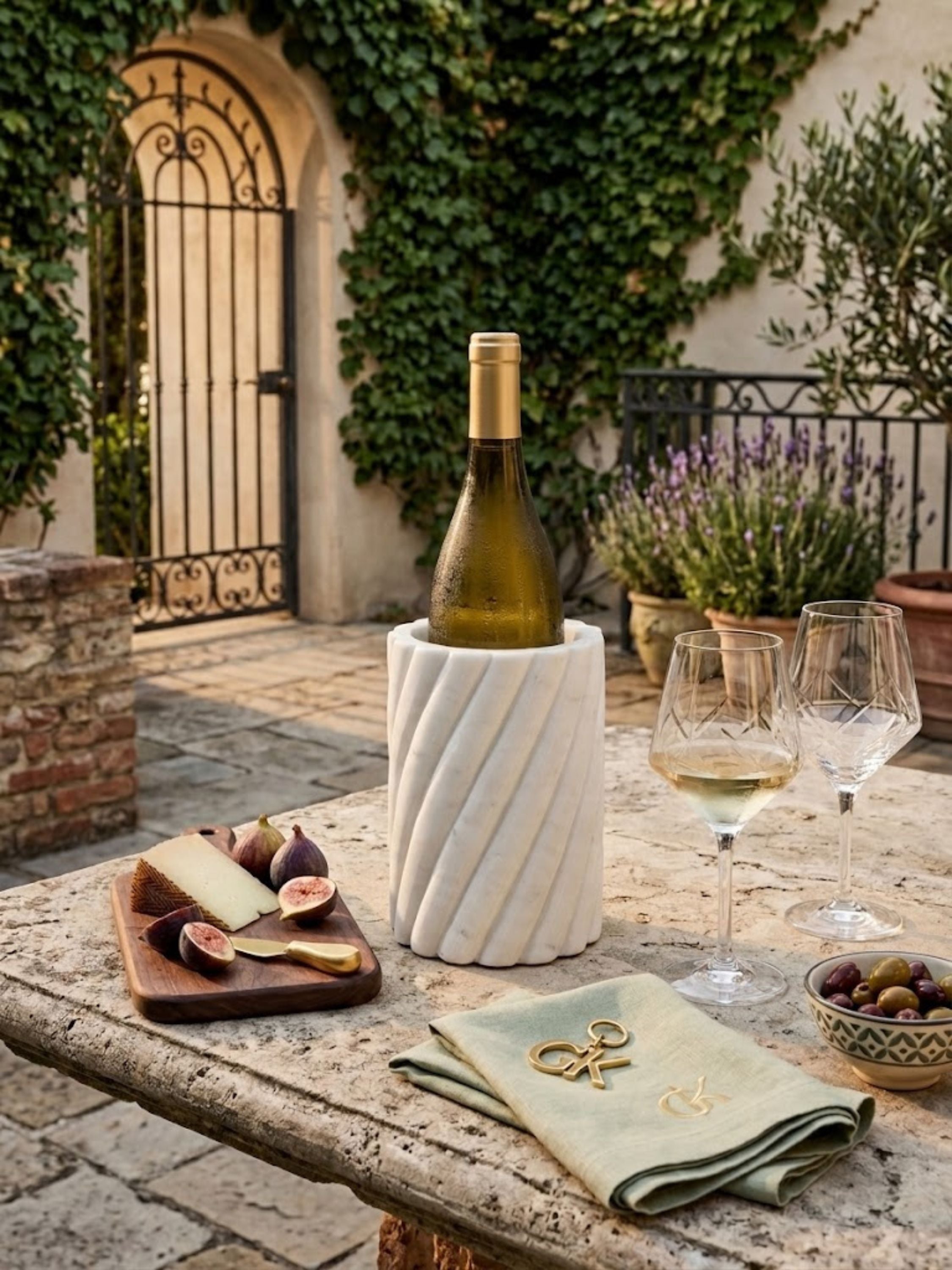 White marble wine cooler with fluted carved surface holding a wine bottle, placed on an outdoor stone table with glasses and serving elements