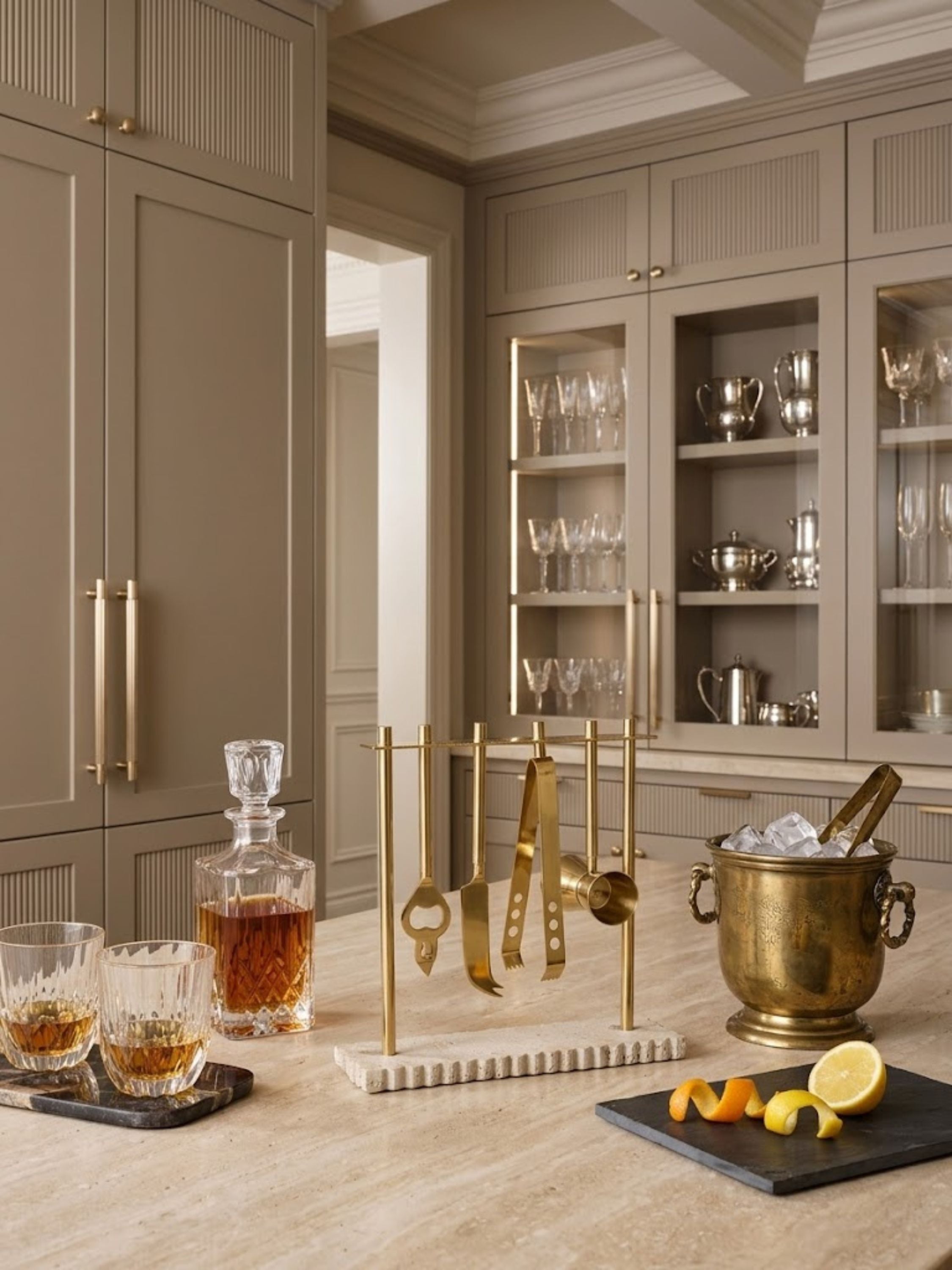 Jubel brass bar tools set displayed on a carved marble stand with cocktail accessories, crystal whiskey glasses, and an ice bucket in a refined home bar setting.