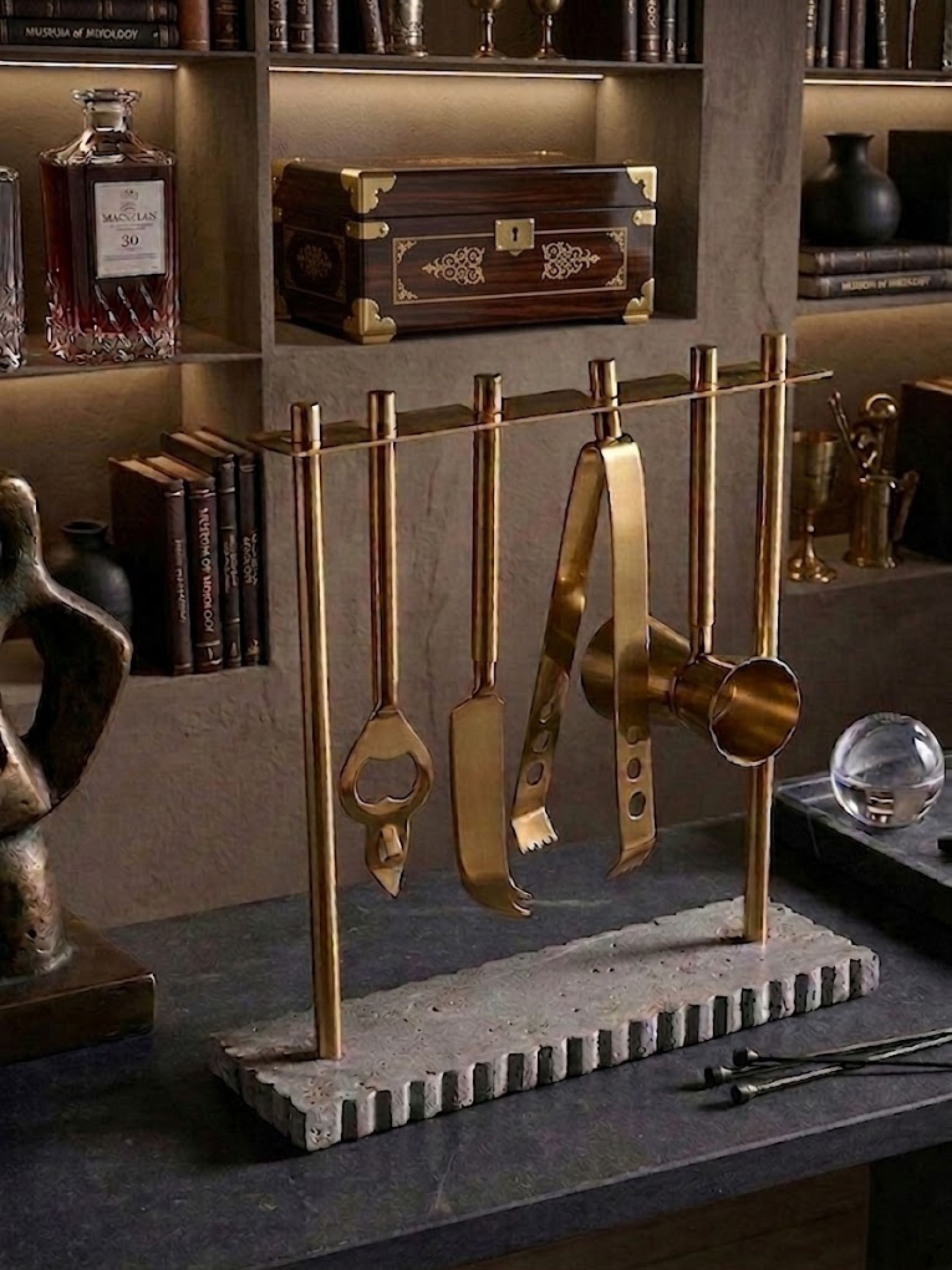Luxury brass bar tools suspended from a marble base stand, featuring a bottle opener, ice tongs, muddler, strainer, and jigger against a sophisticated library bar backdrop.
