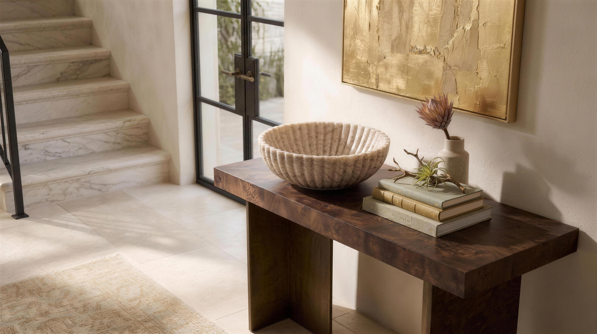 Beige onyx fluted bowl on a burl wood console in an entryway, a pale stone stair and a glazed door behind it