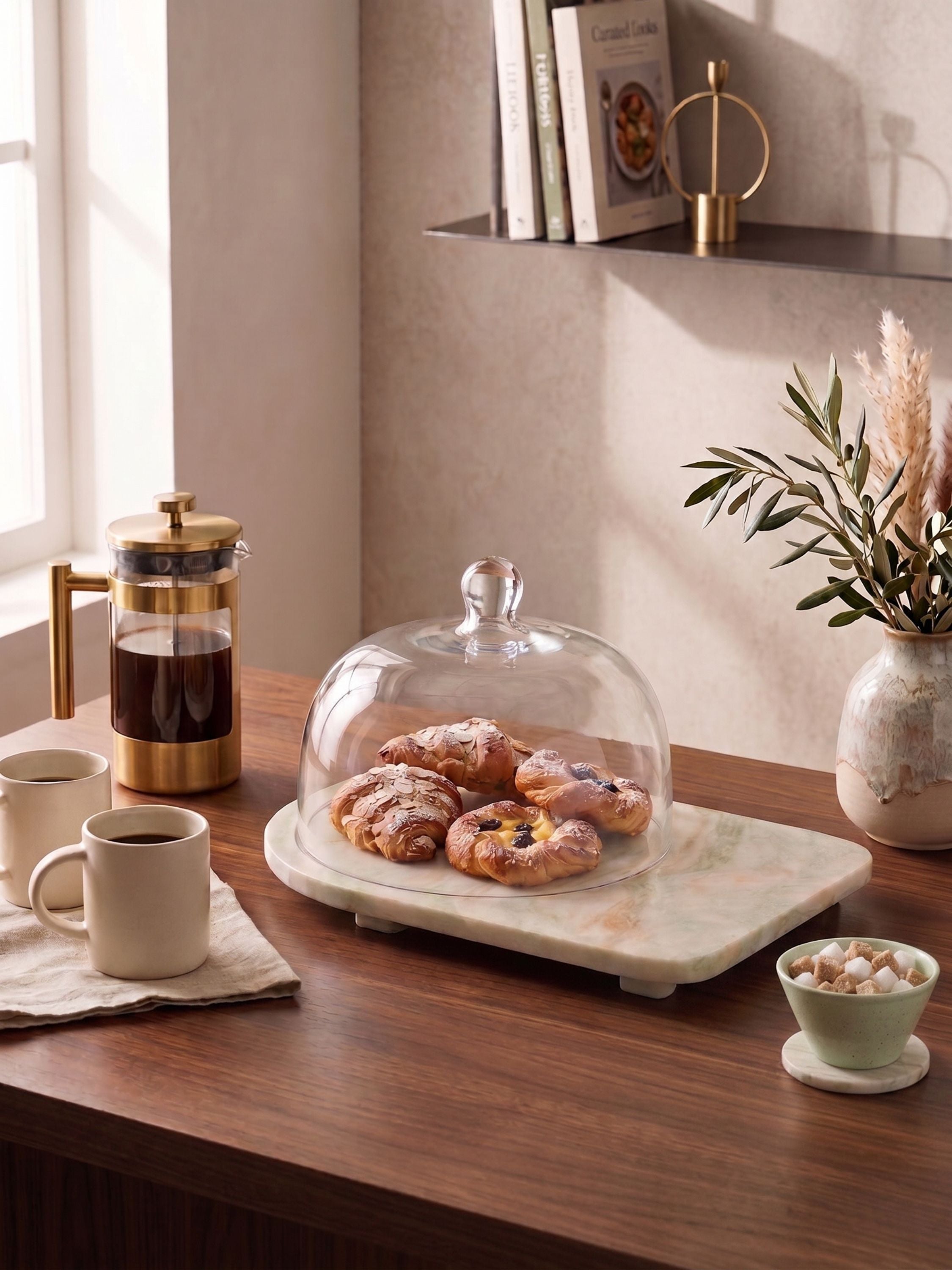 Onyx serving board with glass cloche displaying pastries on a wooden table in a warm kitchen setting
