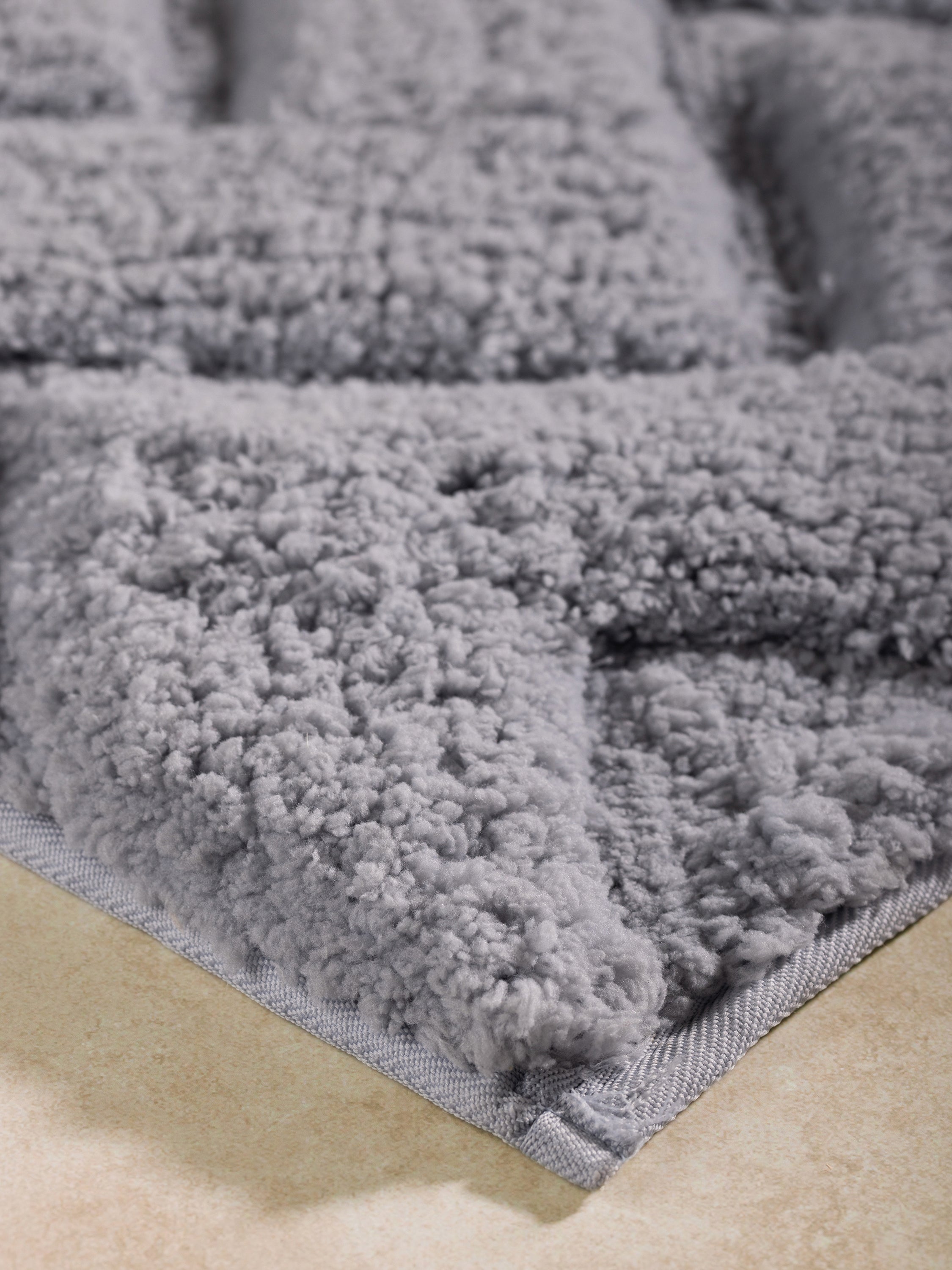 Close-up corner of gray microfiber bath mat, showing dense chevron pile and stitched edge.