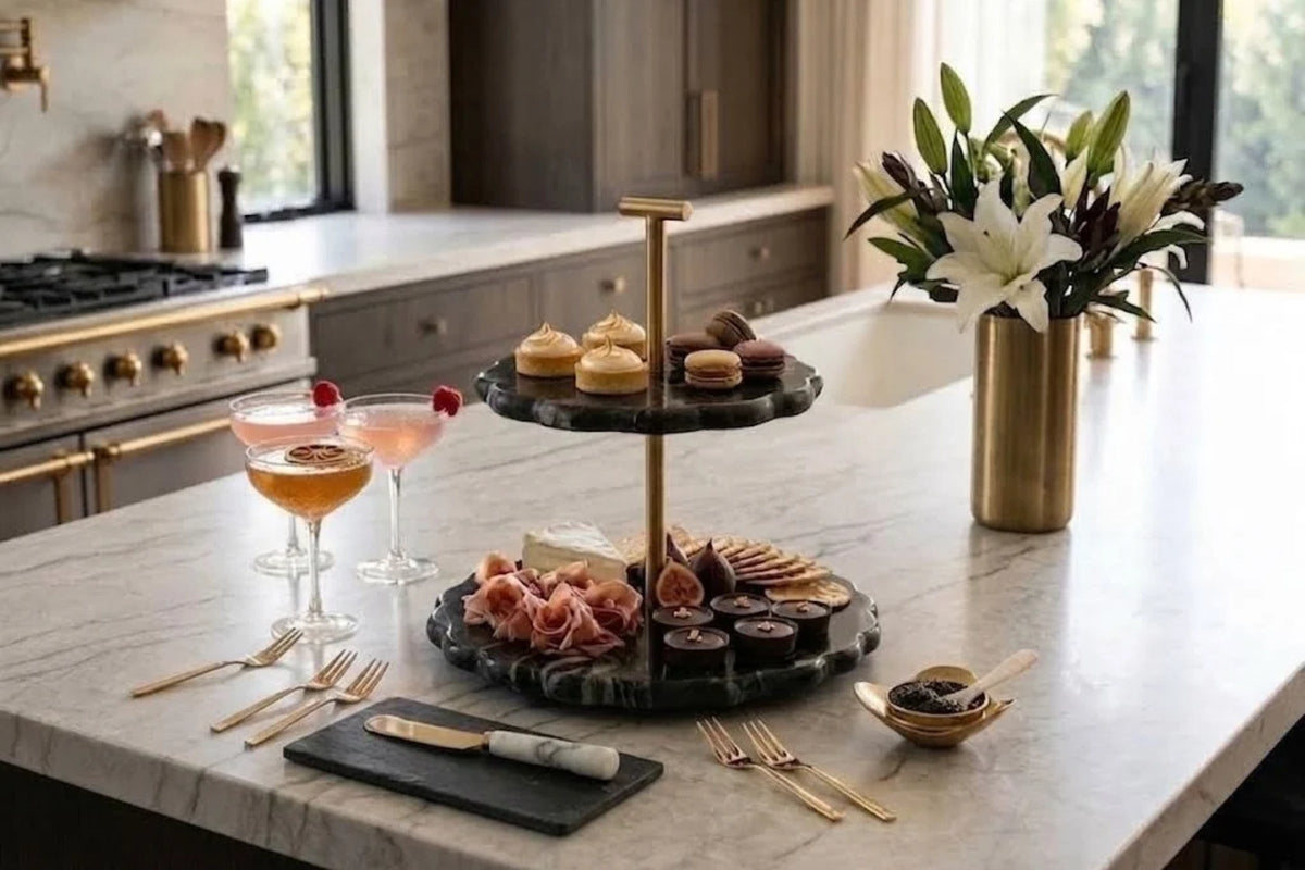 Black marble two-tier stand on a brass stem, set with pastries and charcuterie on a white marble kitchen island