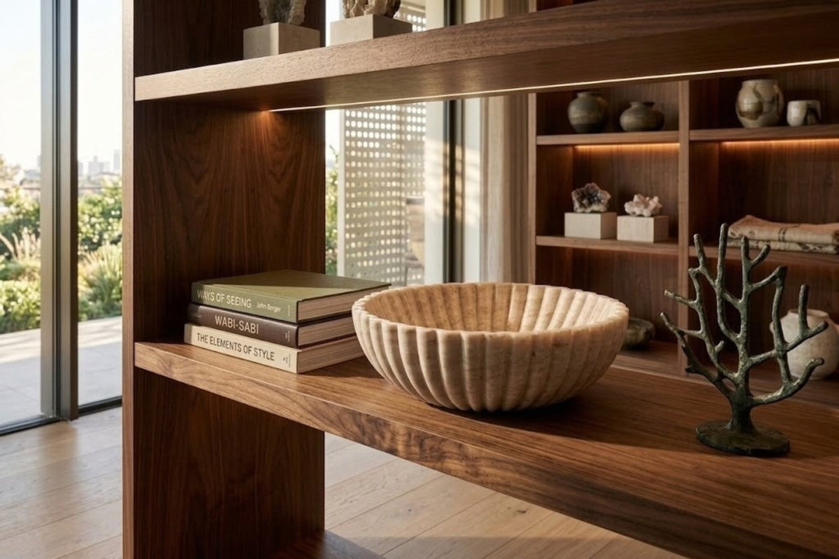 Fluted beige onyx bowl resting empty on a walnut display shelf, low daylight raking across the wood grain