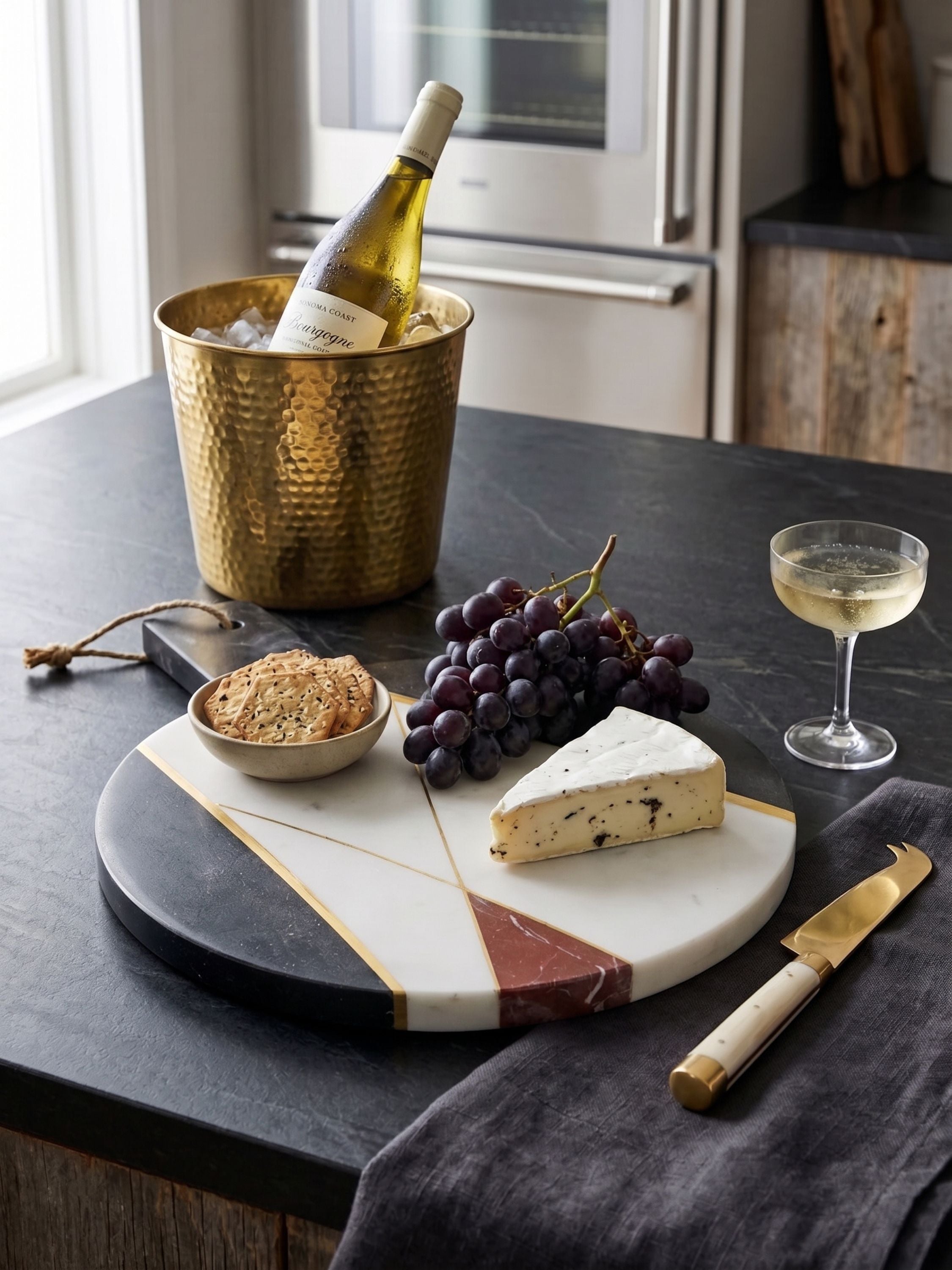 Marble cheese board styled with cheese, grapes, and crackers on a kitchen countertop with wine and barware in a refined serving setting