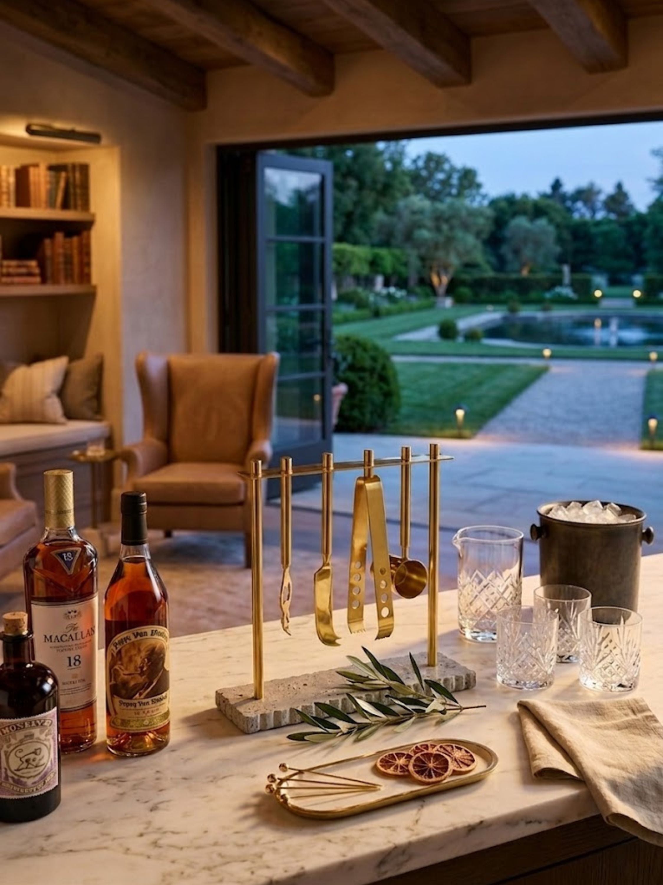 Jubel travertine bar tools styled in a home library, their brushed brass stand set beside whiskey service pieces.