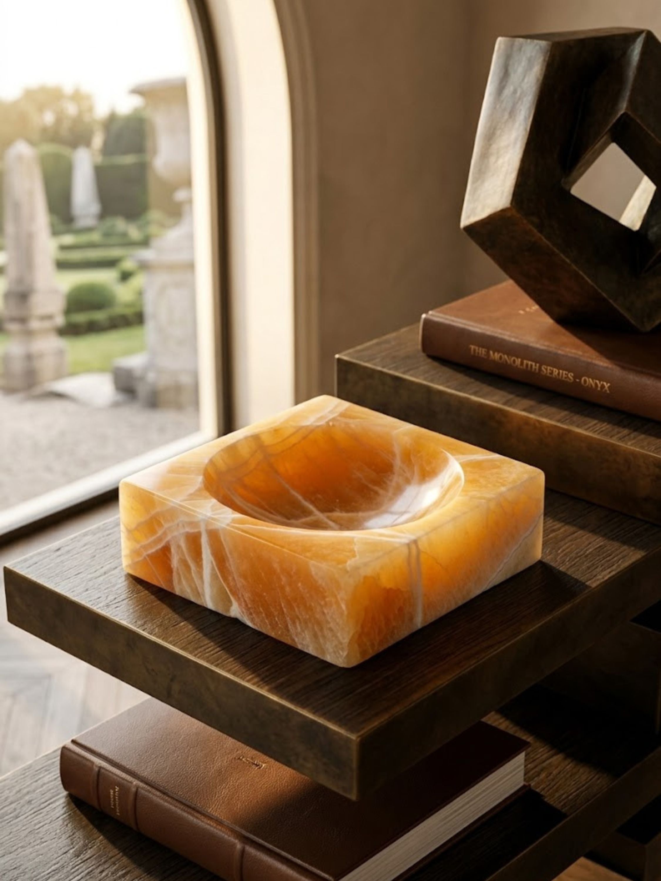 Amber onyx catchall bowl carved from richly veined stone, styled as a sculptural tabletop accent alongside design books and warm walnut finishes.