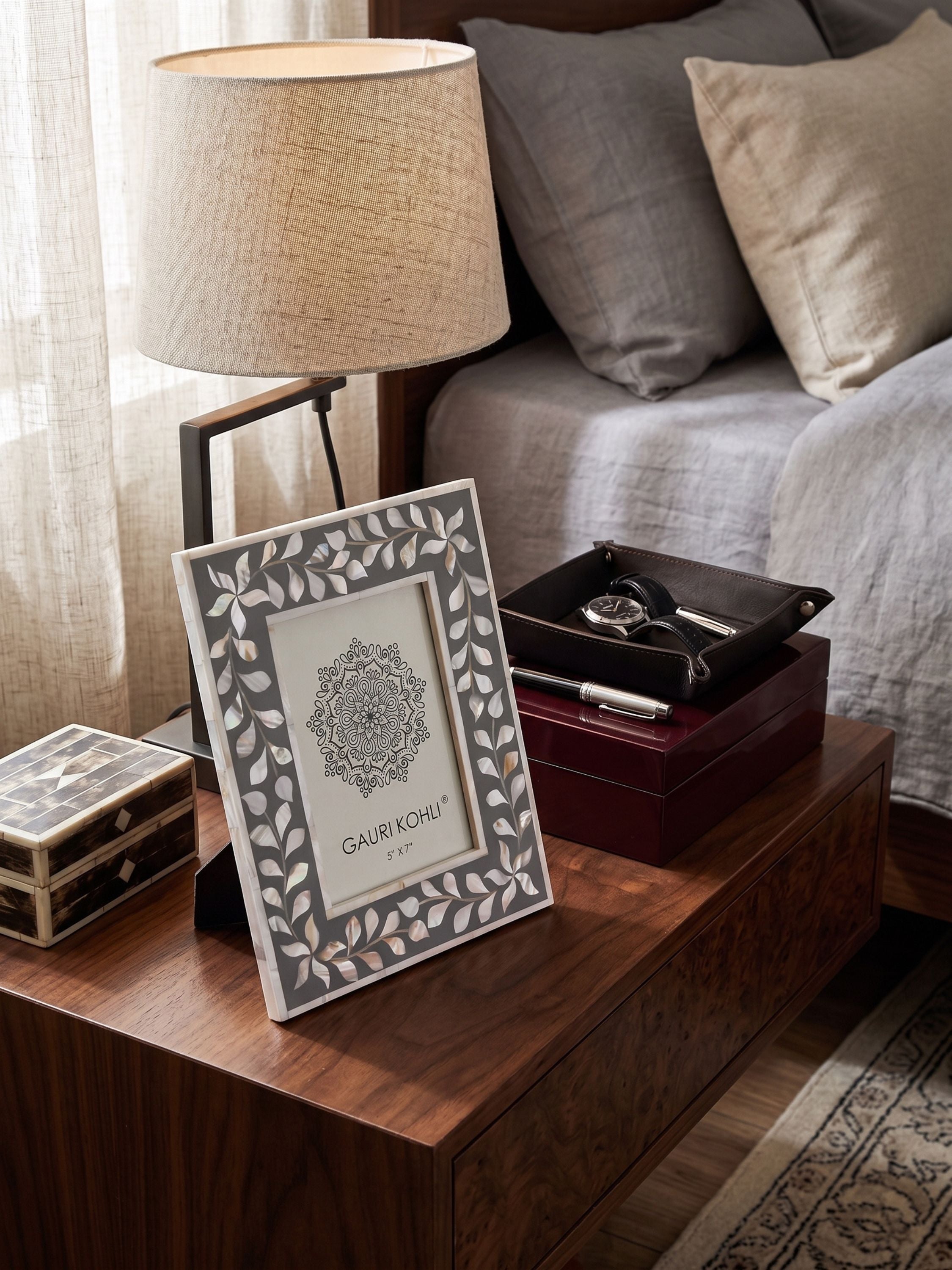 Mother of pearl and grey inlay tabletop photo frame with leaf border design styled on a wooden bedside table with lamp and decor accents