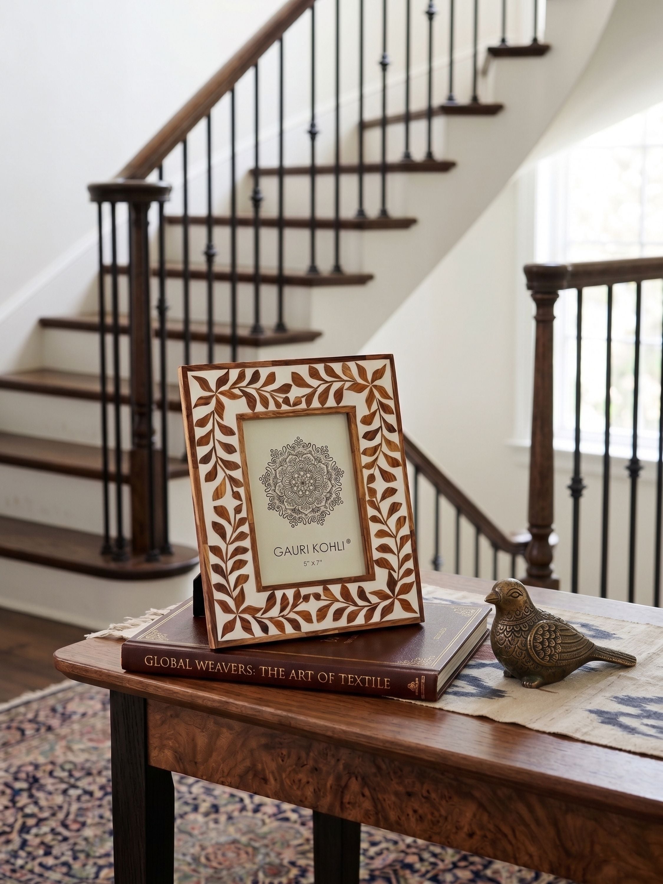 Wood inlay ivory tabletop photo frame with leaf border design styled on a console table with books and decorative bird accent near a staircase
