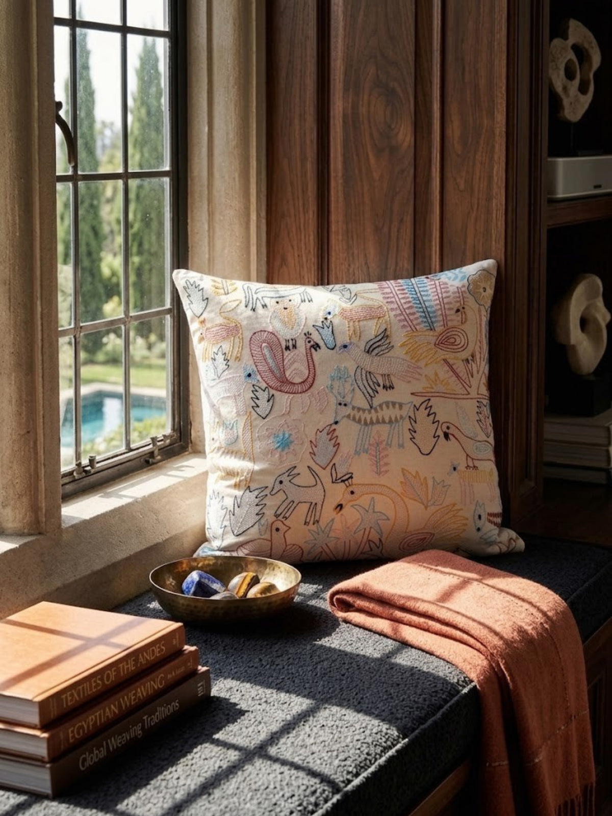 Fateh oat cotton pillow with fine outlined figures, on a dark window seat beside a terracotta throw.