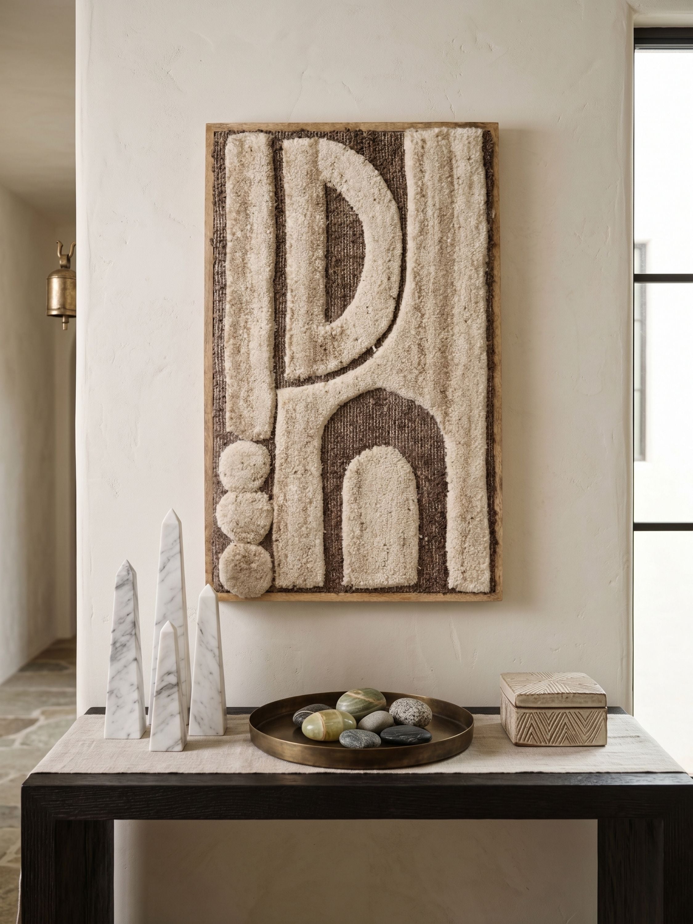 Ivory and brown tufted wool wall art with abstract arch forms and raised texture, placed above a console with decorative stone objects in a warm interior