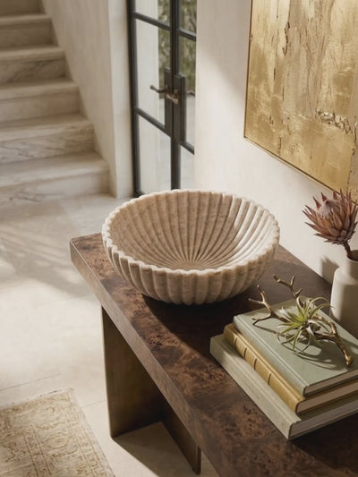 Beige onyx fluted bowl on a burl wood console beneath a gilt canvas, the camera easing slowly toward it as afternoon light moves on the stone stair